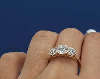 May include: A gold ring with three clear, round gemstones. The ring is worn on a finger, set against a blue backdrop. The gemstones are the main feature, highlighting their sparkle and clarity.