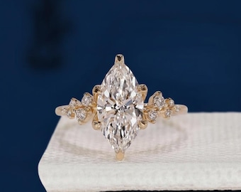 Könnte beinhalten: Ein goldener Verlobungsring mit einem großen Diamanten im Marquiseschliff als Mittelpunkt. Der Ring hat kleinere Diamanten auf beiden Seiten des Hauptsteins, auf einer weißen Oberfläche vor einem dunkelblauen Hintergrund.