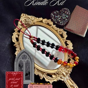 Op de afbeelding: Een handspiegel met een gouden frame, een kralenketting, een hartvormig object en een Kindle-hoesje. De tekst "Moody Dragon Rider Kindle Kit" is zichtbaar. Extra kaarten lezen "girls just want to ride dragons" en "Dragons & Smut Book Club".