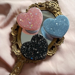 Peut inclure: Trois supports de téléphone en forme de cœur avec des paillettes. Un rose, un bleu et un noir. Ils sont disposés sur un miroir doré avec une poignée ornée. Le fond est un tissu rose pâle.