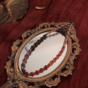 May include: A beaded necklace with a gradient of colors, from deep burgundy to orange, is reflected in a vintage hand mirror. The mirror has an ornate gold frame, and the necklace is displayed on a red fabric background.