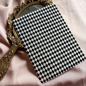 Peut inclure: Une carte à motif de damier noir et blanc est présentée à côté d'un miroir vintage à cadre doré. Le miroir et la carte reposent sur un tissu rose doux, créant un contraste visuel. Le motif de la carte est un motif répétitif de carrés noirs et blancs.