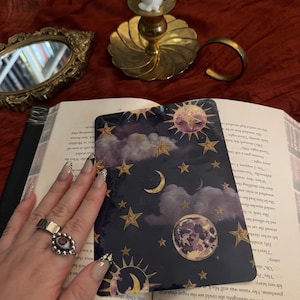 May include: A dark blue card with a celestial design featuring gold stars, moons, and suns. The card is held over an open book. A silver ring with an eye design is visible. A gold candle holder and a small mirror are in the background.