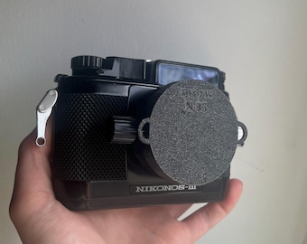 May include: A black Nikonos-III camera with a lens cap. The camera has a textured grip and a silver lever on the side. The lens cap has the text "FISHEYE N 35".