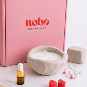 May include: A pink box with the text nobo NOT BORING DIY SETS, a beige textured bowl candle, a small amber glass dropper bottle with a white cap, two red heart-shaped wax pieces, a beige block, and several small white and pink letter beads.