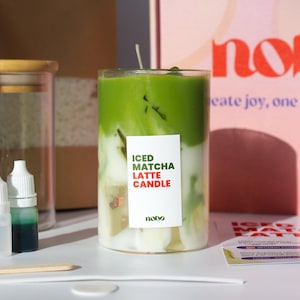 Könnte beinhalten: Eine geschichtete Kerze in einem klaren Glas mit grünem und weißem Wachs, beschriftet mit "Iced Matcha Latte Candle". Das Bild enthält kleine Flaschen, einen Holzstab und ein Glas mit Holzdeckel. Ein rosa und roter Hintergrund zeigt das Wort "nobo".