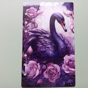Könnte beinhalten: Planereinlage mit einer Aquarellillustration eines schwarzen Schwans mit rotem Schnabel, umgeben von lila Rosen und Blättern. Der Schwan hat einen gebogenen Hals vor einem weichen, lila Hintergrund. Die Einlage hat vorgestanzte Löcher.