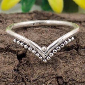 Puede incluir: Un anillo de plata en forma de cheurón con una fila de pequeñas cuentas redondas a lo largo de la forma de V. El anillo está sobre una superficie marrón texturizada, con follaje verde y amarillo en el fondo. Es una joya.