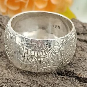 Op de afbeelding: Een brede, zilveren ring met een gegraveerd bloemmotief. De ring is aan de binnenkant gemarkeerd met "925". De ring staat tegen een getextureerde, bruine achtergrond.