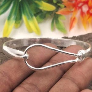 May include: A silver-colored bangle bracelet with a unique hook-and-eye closure. The bracelet is round and has a smooth, polished finish. The hook closure is a teardrop shape. The bracelet is shown against a blurred background of green and orange.