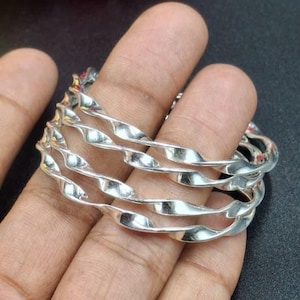 Könnte beinhalten: Ein Satz silberfarbener Armreifen mit einem gedrehten Seildesign. Die Armreifen sind übereinander gestapelt und reflektieren das Licht. Der Schmuck wird vor einem dunklen Hintergrund präsentiert.