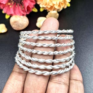 Può includere: Un braccialetto color argento con un design a corda intrecciata. Il braccialetto è avvolto attorno a un dito, mettendo in risalto la sua trama intricata e la lucentezza metallica. Il gioiello è ambientato su uno sfondo scuro con fiori.