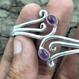 May include: A silver-toned bracelet with an open design, featuring two oval, purple gemstones. The bracelet has a decorative, swirling pattern and is held by a hand.