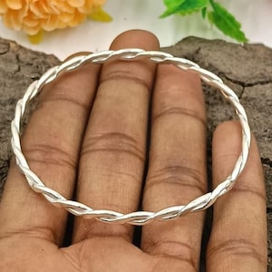 Può includere: Un braccialetto rigido color argento con un motivo a corda intrecciata. Il braccialetto rotondo sembra essere in metallo. Il braccialetto è tenuto in mano, con uno sfondo sfocato di fiori e legno.
