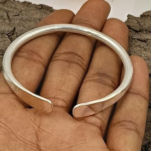 May include: A silver-colored, open cuff bracelet with a smooth, rounded design. The bracelet is shown on a hand, with the open ends facing the viewer. The jewelry is likely made of metal and has a polished finish.