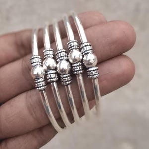 Può includere: Un set di cinque braccialetti d'argento. Ogni braccialetto presenta una serie di perline decorative e una perlina centrale più grande. I braccialetti sono esposti su uno sfondo neutro, evidenziando la loro finitura argentata lucida. Un accessorio elegante.