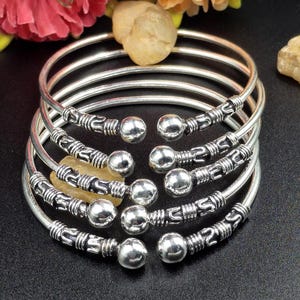 Op de afbeelding: Een stapel zilverkleurige armbanden met decoratieve details en ronde kogelaccenten. De armbanden zijn in een halve cirkel gerangschikt, met een donkere achtergrond en een paar decoratieve elementen. De diameter van elke armband is ongeveer 6 cm.