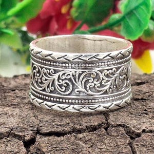 Può includere: Un anello largo in argento con intricati disegni floreali e a volute. L'anello presenta motivi e texture dettagliati, con una fascia di piccole perle e una serie di forme a freccia. L'anello è posto su uno sfondo di legno naturale.