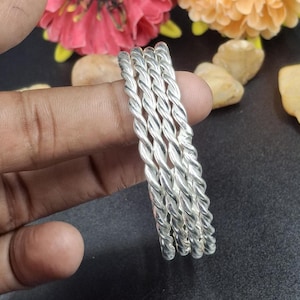 Puede incluir: Un conjunto de cinco pulseras plateadas con diseño de cuerda trenzada. Las pulseras se sostienen sobre un fondo oscuro con flores y rocas. Las pulseras de metal brillante tienen una superficie texturizada. Cada pulsera mide aproximadamente 0,5 cm de grosor.