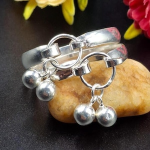 May include: Close-up of two silver anklets with dangling silver bells. The anklets are resting on a light brown stone, with a dark background. The anklets have a simple, elegant design, perfect for adding a touch of sparkle to any outfit.