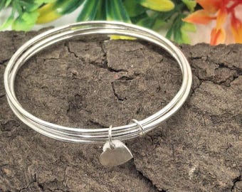 Pode incluir: Um conjunto de pulseiras de prata com um pequeno pingente de coração. As pulseiras são empilhadas e repousam sobre uma superfície texturizada marrom escura. O pingente de coração está pendurado.