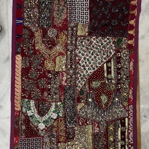 May include: A rectangular wall hanging featuring a patchwork of deep red fabric panels. The surface is heavily embellished with intricate gold embroidery, metallic sequins, small mirrors, and beadwork in various floral and geometric patterns.
