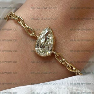 May include: A gold chain bracelet featuring a large, pear-shaped diamond. The diamond is the focal point, set in a gold bezel, and connected to the chain. The bracelet is displayed on a person's wrist, against a white shirt.