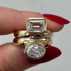 May include: Three gold rings with clear gemstones. The top ring features an emerald-cut stone, the middle ring is a plain band, and the bottom ring has a round-cut stone. The rings are held up by a person with red nail polish.
