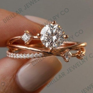 May include: A rose gold ring featuring a large round brilliant-cut center stone, two smaller princess-cut side stones, and a crossover band design. One section of the band is pavé-set with small round stones, while the other is smooth metal.