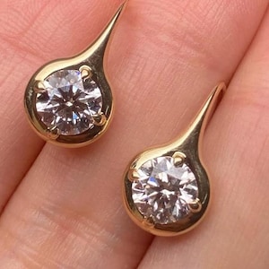 May include: A pair of gold earrings, each featuring a round, clear gemstone set in a teardrop-shaped gold setting. The earrings are held in a person's hand, showcasing their design and sparkle. The earrings have a post and are designed to be worn in pierced ears.