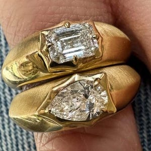 Peut inclure: Deux bagues en or avec de grosses pierres précieuses claires. L'une présente un diamant taille émeraude, l'autre un diamant en forme de poire. Les bagues ont une finition en or brossé et sont portées sur un doigt.
