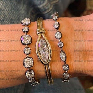 May include: A close-up of three pieces of jewellery. Two bracelets with square and round diamonds and a gold bracelet with a large marquise-cut diamond. The bracelets are on a wrist. The text "BRILLAR JEWELRY CO" is visible.