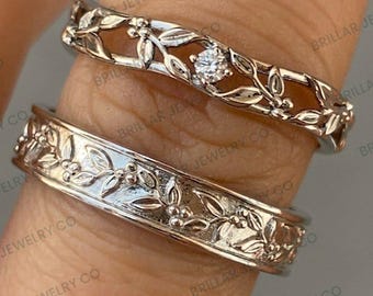 Puede incluir: Dos anillos de tono plateado con un motivo botánico de hojas y pequeñas bayas. El anillo superior es una banda calada con una gema redonda transparente engastada en el centro. El anillo inferior es una banda sólida con un patrón de hojas en relieve.