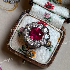 Puede incluir: Un anillo de estilo vintage con una gran gema roja central, rodeada de piedras más pequeñas y claras. El anillo se muestra en un joyero antiguo forrado con un estampado floral, junto con un boceto del anillo. El texto "BrillarjewelryCo" es visible.