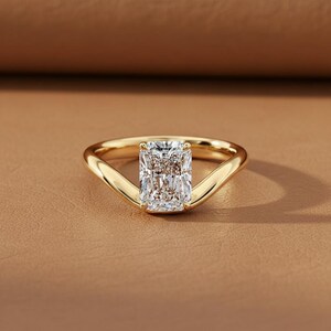 May include: A radiant-cut diamond ring with a gold band. The diamond is rectangular and set in a gold band that curves up to meet the diamond. The ring is displayed on a brown surface.
