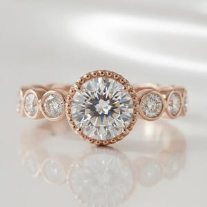 May include: Rose gold ring featuring a large, round, clear gemstone at the center, surrounded by a beaded halo. Smaller, clear gemstones are set along the band. The ring is displayed on a reflective surface.