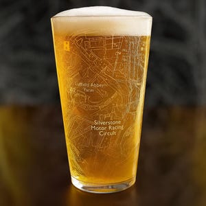 May include: A pint glass filled with beer, featuring a foamy head. The glass is etched with a map of the Silverstone Motor Racing Circuit and Luffield Abbey Farm. The beer is a golden colour.