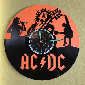 Könnte beinhalten: Eine schwarz-orangefarbene Wanduhr im AC/DC-Band-Design. Das Zifferblatt ist eine Schallplatte mit Silhouetten von Bandmitgliedern. Die Zeiger der Uhr sind grün. Das Logo der Band ist abgebildet.