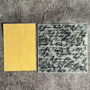 May include: A rectangular yellow card with an embossed cursive script pattern and a square translucent plastic embossing folder featuring a detailed black printed script design that mimics handwritten calligraphy across the entire surface.