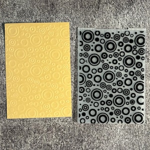 May include: A rectangular yellow card with an embossed pattern of concentric circles and a rectangular translucent plastic embossing folder featuring a black printed pattern of various sized concentric circles and dots.