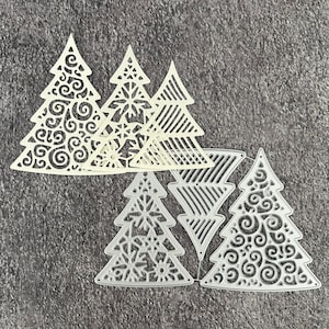 May include: A collection of die-cut Christmas tree shapes in off-white and silver. The trees feature intricate designs, including swirls, snowflakes, and linear patterns. The shapes are arranged on a textured gray surface, suggesting a craft or DIY project.