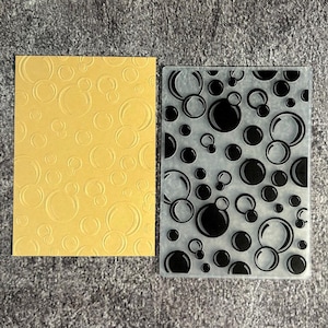 May include: A rectangular yellow cardstock sheet featuring an embossed bubble pattern alongside a clear plastic embossing folder with a matching black and white printed bubble design, both displaying a variety of circular shapes in different sizes.