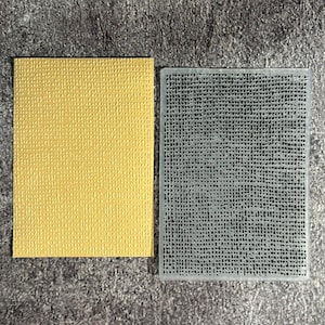 May include: A rectangular yellow cardstock sheet with a raised, grid-like texture next to a clear, rectangular plastic embossing folder featuring a matching recessed cross-hatch pattern designed to create impressions on paper surfaces.
