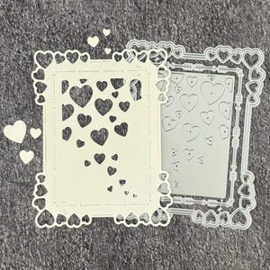 May include: Two die-cut frames with heart-shaped cutouts and borders. One frame is cream-coloured, the other is silver. Several loose heart-shaped cutouts are scattered around the frames. The frames are likely for crafting or scrapbooking.