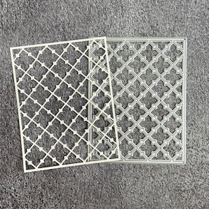 May include: Two white metal die cut stencils with a geometric lattice pattern. One stencil is fully cut, and the other is partially cut. The stencils are rectangular and rest on a textured gray surface.