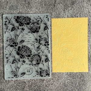May include: A rectangular translucent plastic embossing folder featuring a floral pattern of peonies and script text, alongside a yellow cardstock sheet embossed with the same design, showing the raised floral and text details clearly.