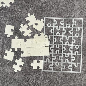 May include: A collection of white puzzle pieces scattered on a gray surface. A metal die-cut frame of a puzzle is also present. The puzzle pieces are made of a light-colored material.