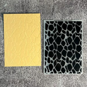 May include: A rectangular yellow card with a raised giraffe-like spot pattern and a clear plastic embossing folder featuring a matching black and white giraffe-like spot design, both displayed side by side on a textured grey surface.