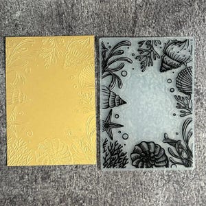 May include: A rectangular clear plastic embossing folder featuring a raised sea-themed pattern of seashells, coral, and bubbles, displayed alongside a gold-coloured cardstock sheet embossed with the same intricate underwater design.