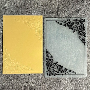 May include: A rectangular gold card featuring an embossed floral corner pattern alongside a clear plastic embossing folder with a matching black printed floral corner design. The set displays a decorative border motif on both the paper and the folder.